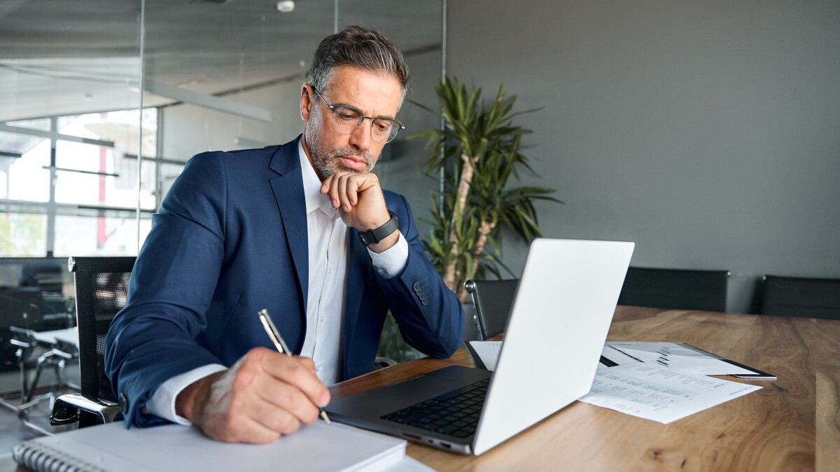 busy middle aged business man sitting at desk working on laptop writing.